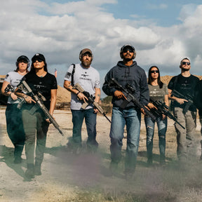 A group of six confidently walks a dirt path, showcasing their 2A Edition firearms. Theyre dressed in casual outdoor clothing and Grunt Styles Come and Take It - Military Green sunglasses. The background features an open sky with clouds, embodying their right to bear arms spirit.