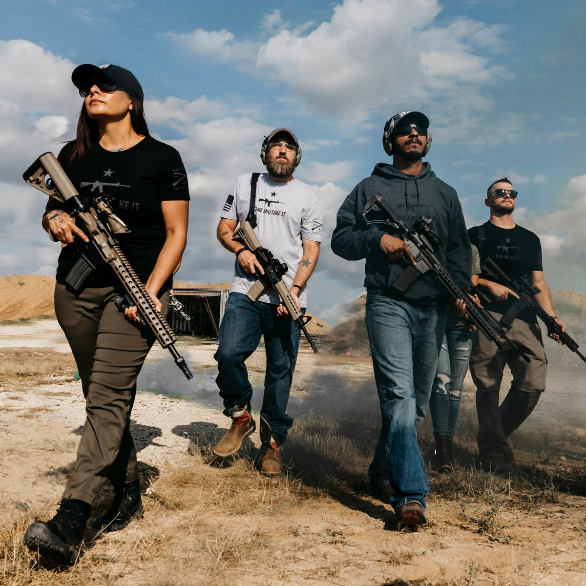 Four individuals, wearing Grunt Styles Come and Take It - Military Green tactical gear, walk a dirt path, focused and purposeful under a partly cloudy sky. The group includes three men and one woman in casual yet tactical attire for this 2A Edition scene.