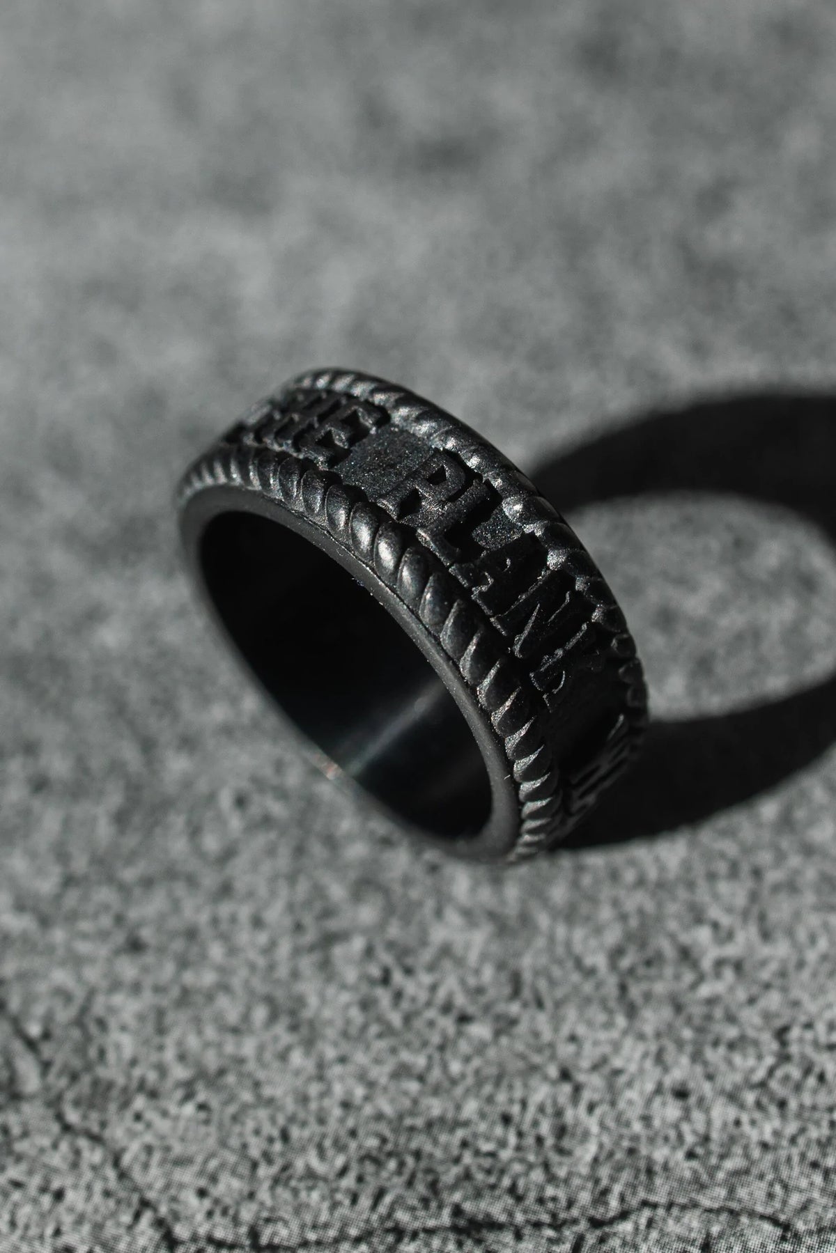 The Walk The Plank - Black Ring by Relentless Betrayal, a dark handcrafted stainless steel ring with a woven texture and engraved letters, rests on a textured gray surface. Its soft shadow adds depth to the image.