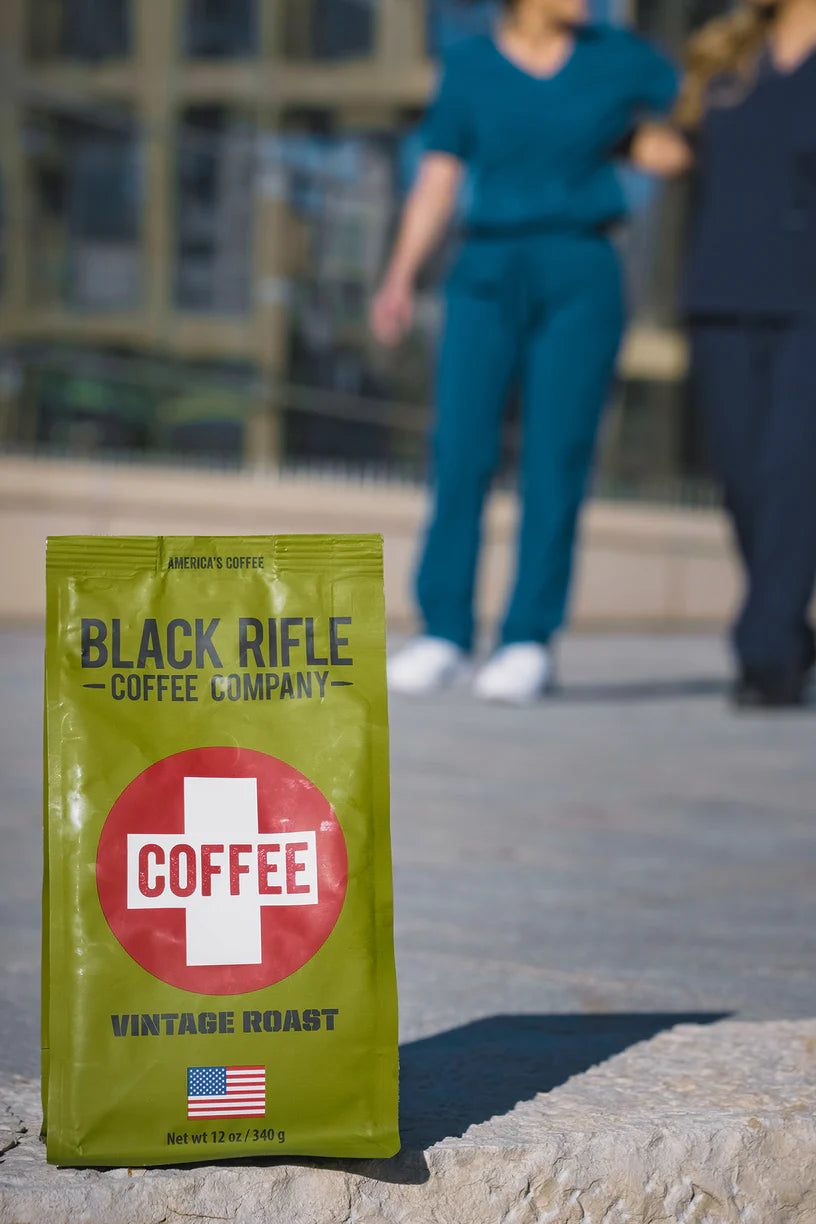 A bag of Black Rifle Coffees Coffee Saves - Whole Bean sits outdoors in the foreground, with two people in medical scrubs blurred in the background.