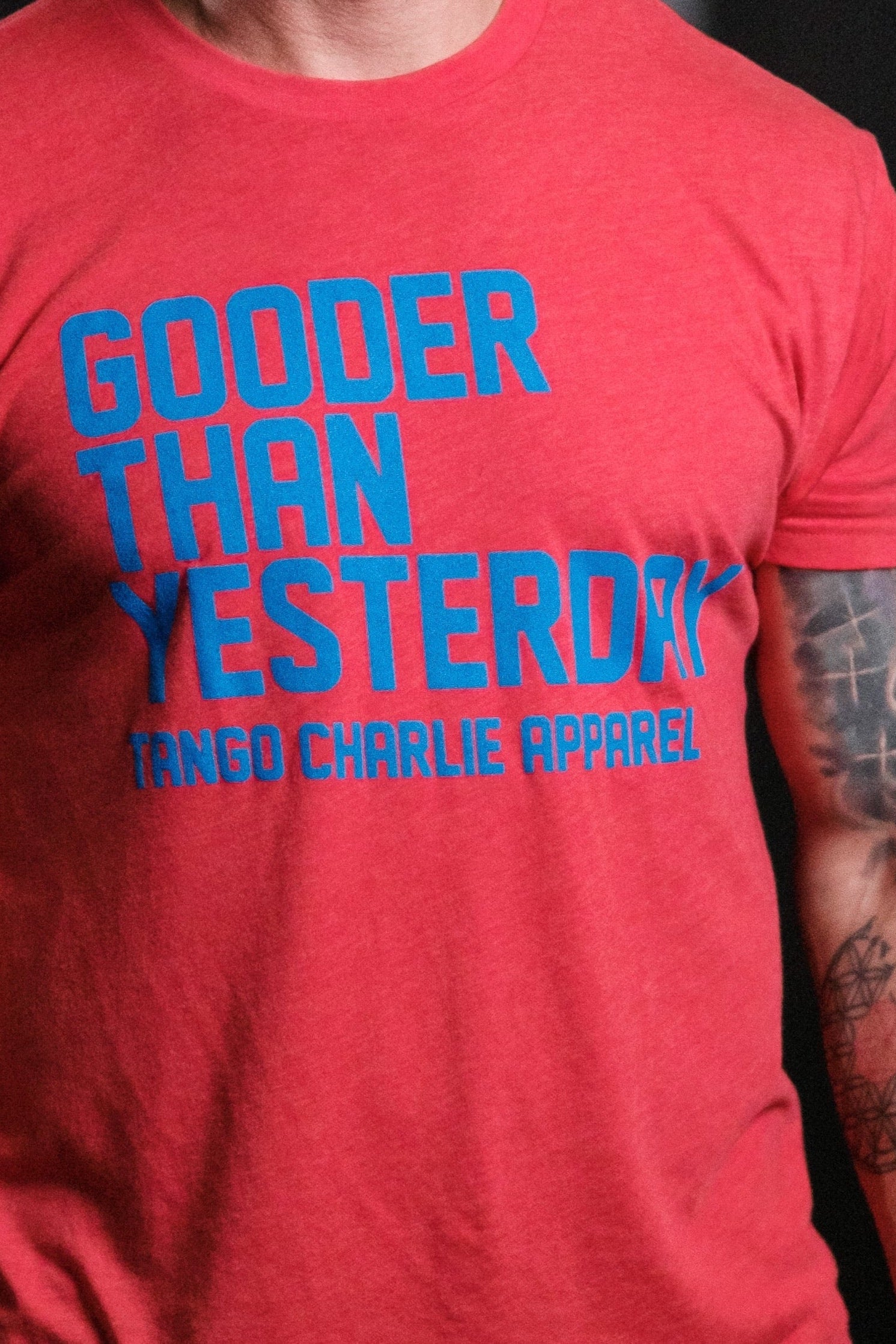 Wearing the cozy red Gooder Than Yesterday T-shirt from Tango Charlie Apparel, featuring blue text, a person with tattooed arms stands out against the black background, highlighting the shirts sleek polyester-cotton blend design.