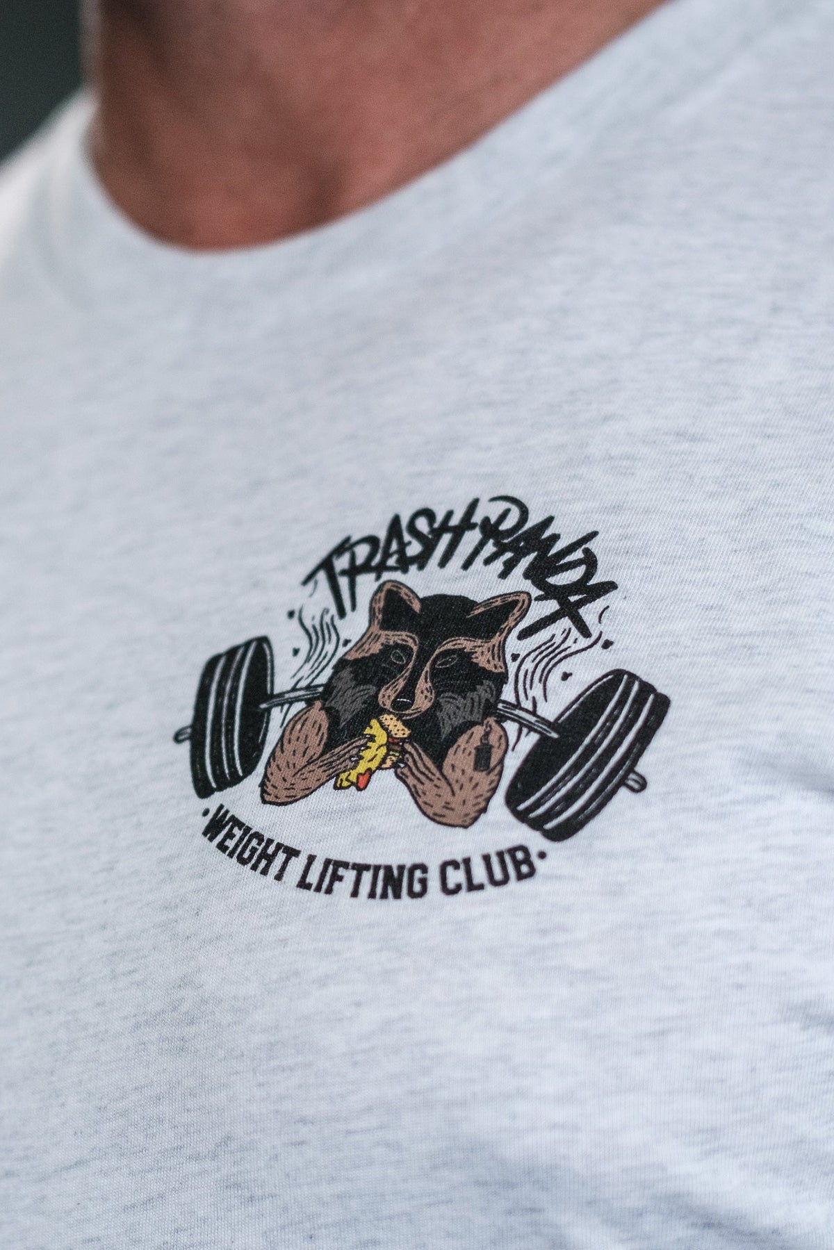 A close-up showcases a person in the Tango Charlie Apparels light gray tri-blend Trash Panda Weight Lifting Club Tee featuring a playful raccoon lifting weights logo encircled by the club’s name, combining strength with style.