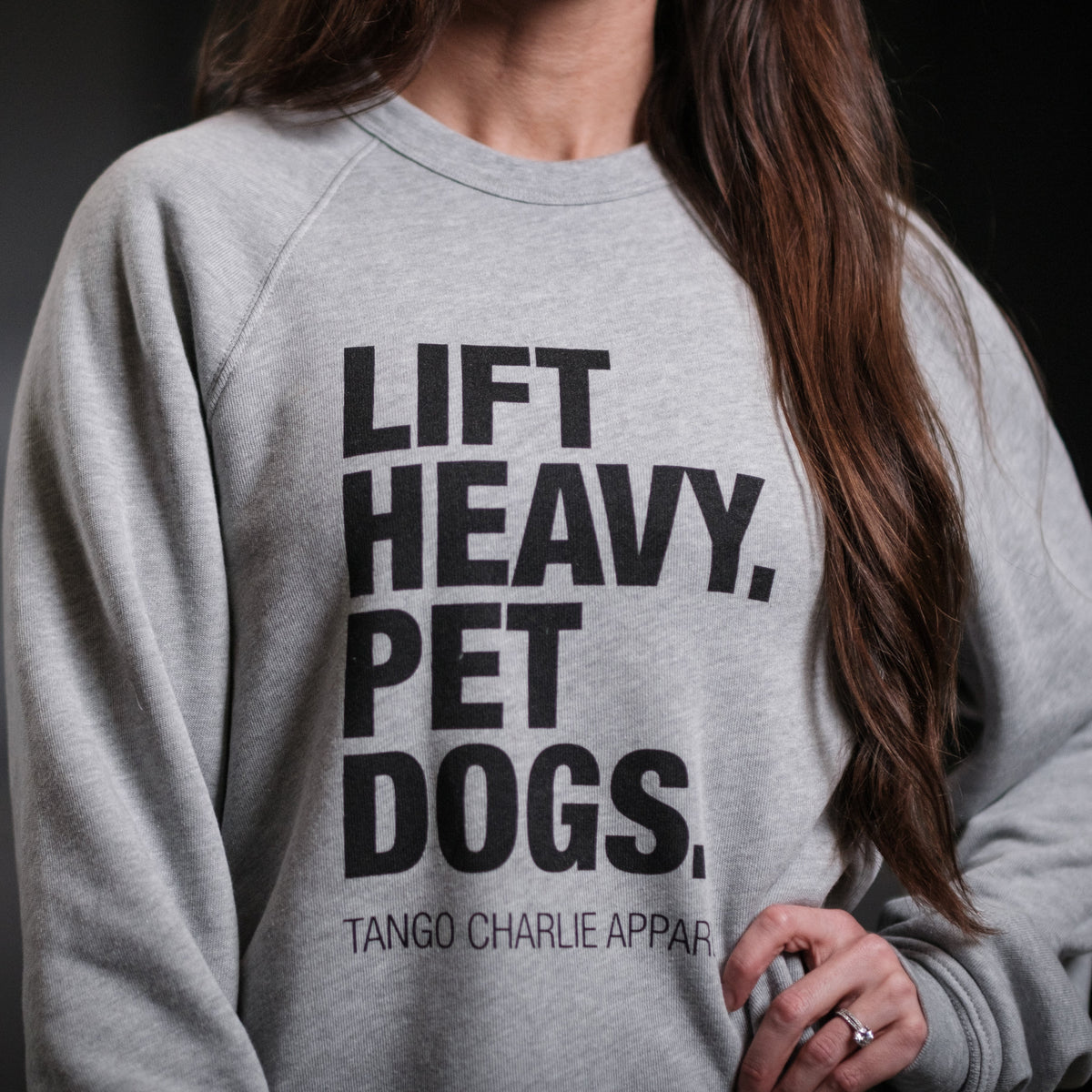 A person wearing the Lift Heavy, Pet Dogs Crewneck by Tango Charlie Apparel showcases LIFT HEAVY. PET DOGS. in bold text, subtly featuring TANGO CHARLIE APPAR. below. Their long brown hair complements their affection for dogs and style by Independent Trading Co.