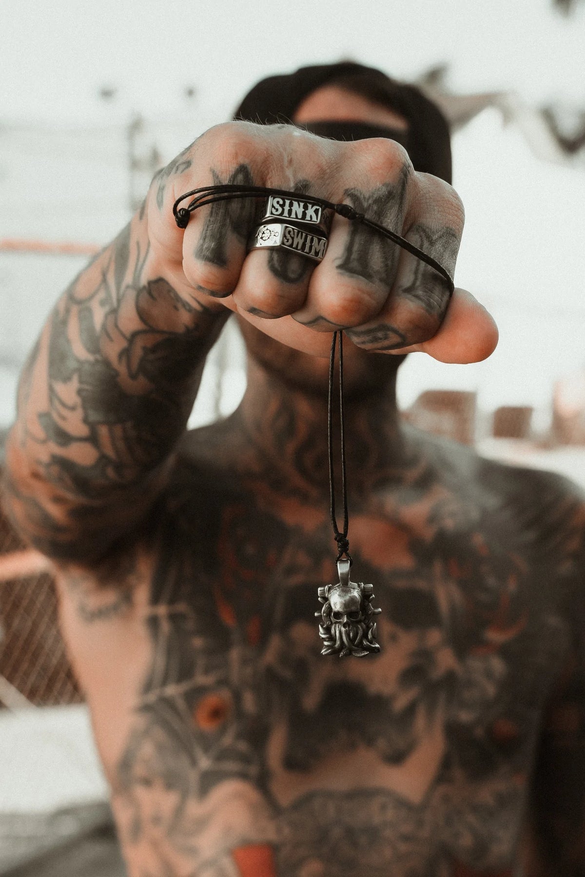 A tattooed person holds out The Kraken Necklace by Relentless Betrayal, featuring a handcrafted pendant. Rings spelling SINS adorn their fingers against a blurred structural backdrop.