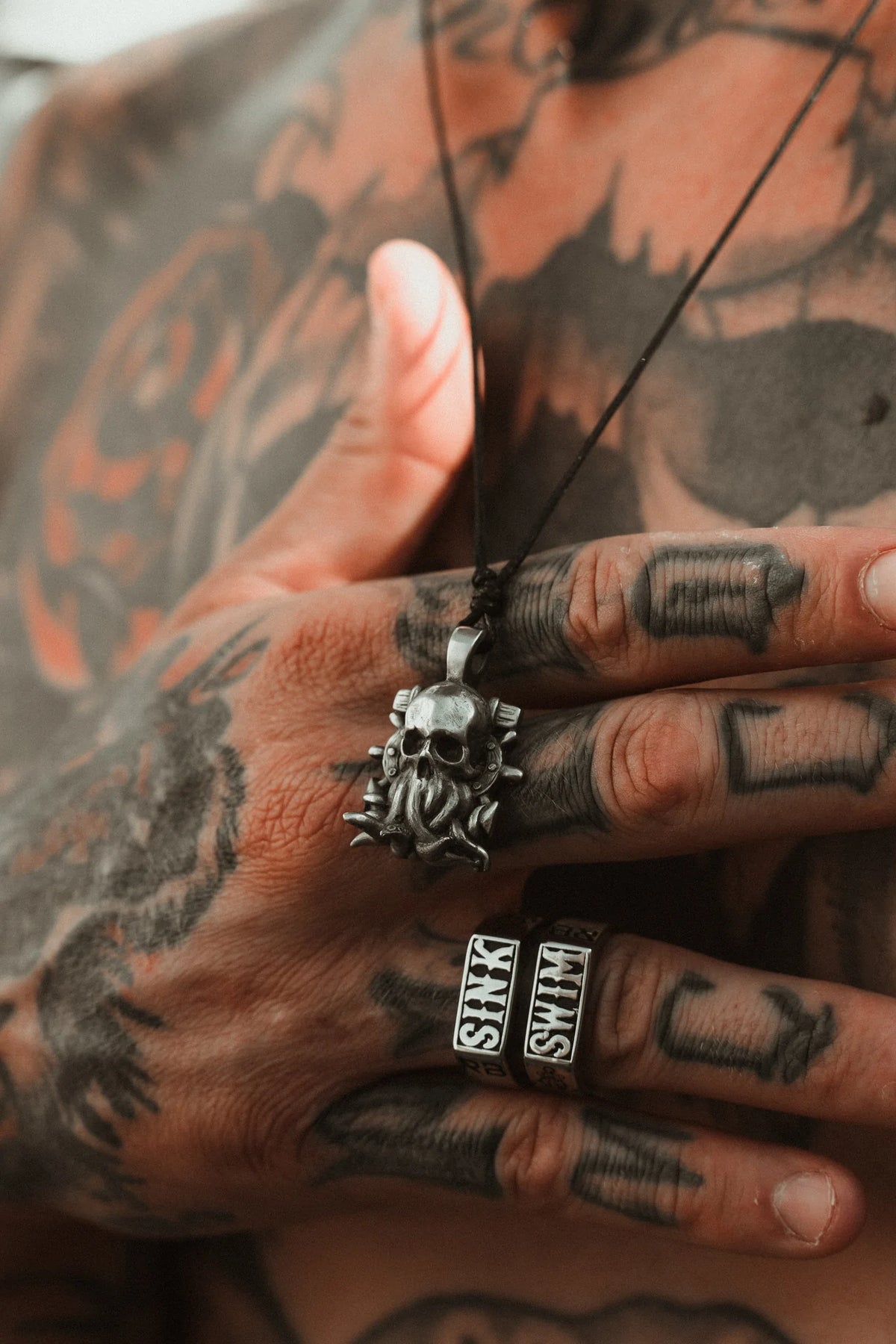 Tattooed hands grip The Kraken Necklace by Relentless Betrayal, a handcrafted pewter pendant with a skull design. The tattooed fingers feature two lettered rings, and the chain suspends the pendant against a chest inked like the mythical krakens embrace.