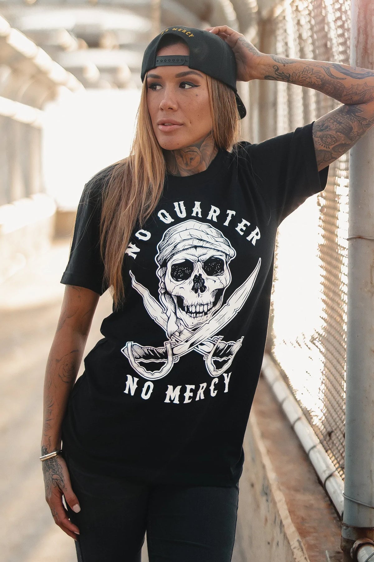 A tattooed individual leans against a chain-link fence on a sunlit path, exuding mercilessness. They wear a backwards black cap and Relentless Betrayals No Quarter Black T-Shirt with a skull, crossed swords, and No Quarter No Mercy emblazoned on it. Their gaze is directed sideways.