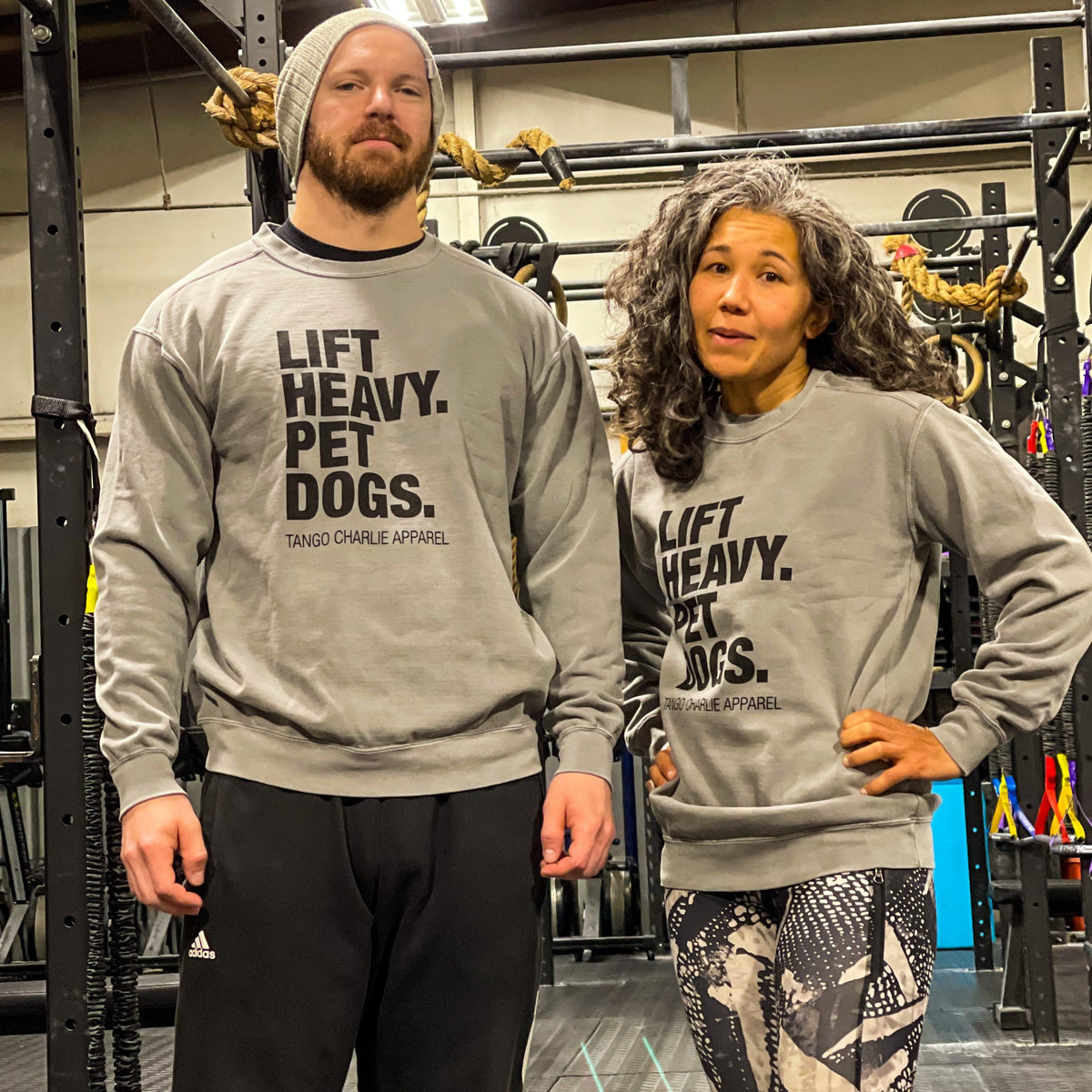 At the gym, a bearded man in a beanie and his curly-haired friend proudly display their love of dogs in matching gray Lift Heavy, Pet Dogs crewnecks from Tango Charlie Apparel amidst ropes and weights.