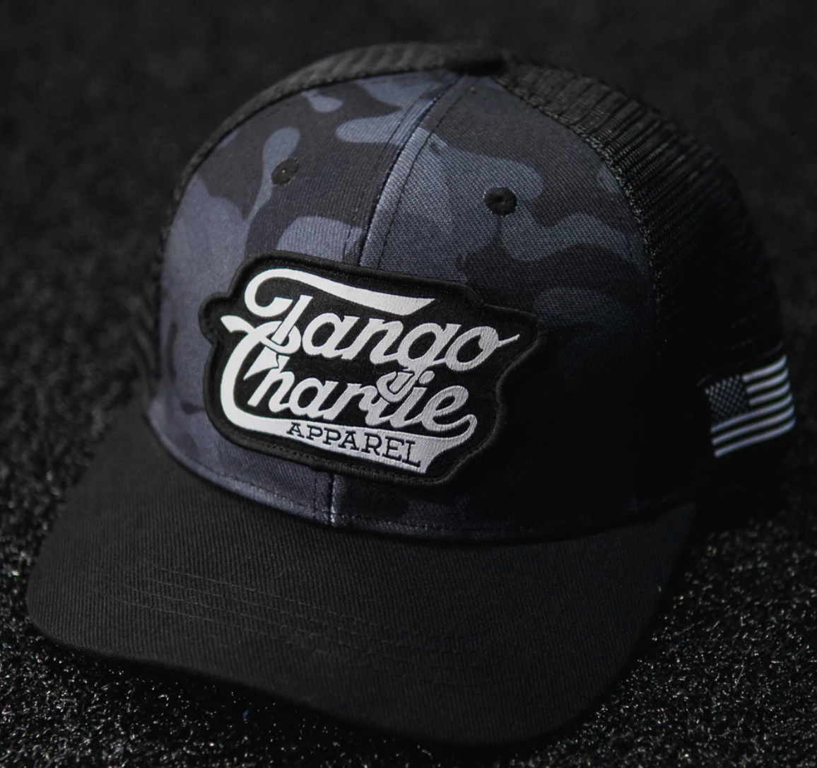 The TCA Black Camo Snapback from Tango Charlie Apparel features a gray camo pattern and black mesh. It displays a Tango Charlie Apparel white script patch on the front and an American flag patch on the mesh, all resting on a textured black surface.