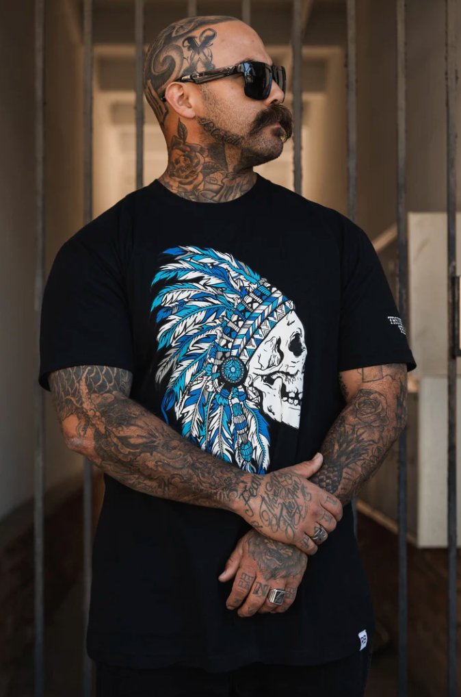 A tattooed man with a mustache and sunglasses stands with arms crossed, wearing the Relentless Betrayal Trustless Chief Tee - Blue, featuring a skull with a blue feathered headdress. The setting of metal bars adds a rebel leader vibe, akin to a prison hallway.