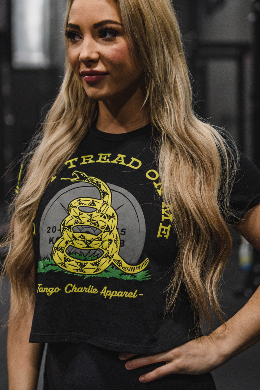 A woman with long blonde hair wears the Don’t Tread On Me Crop Tee by Tango Charlie Apparel—a black, next-level cropped t-shirt crafted from soft polyester cotton and featuring a striking coiled rattlesnake graphic alongside empowering “Don’t Tread On Me” and Tango Charlie Apparel text. This standout design perfectly blends comfort with bold, confident style.