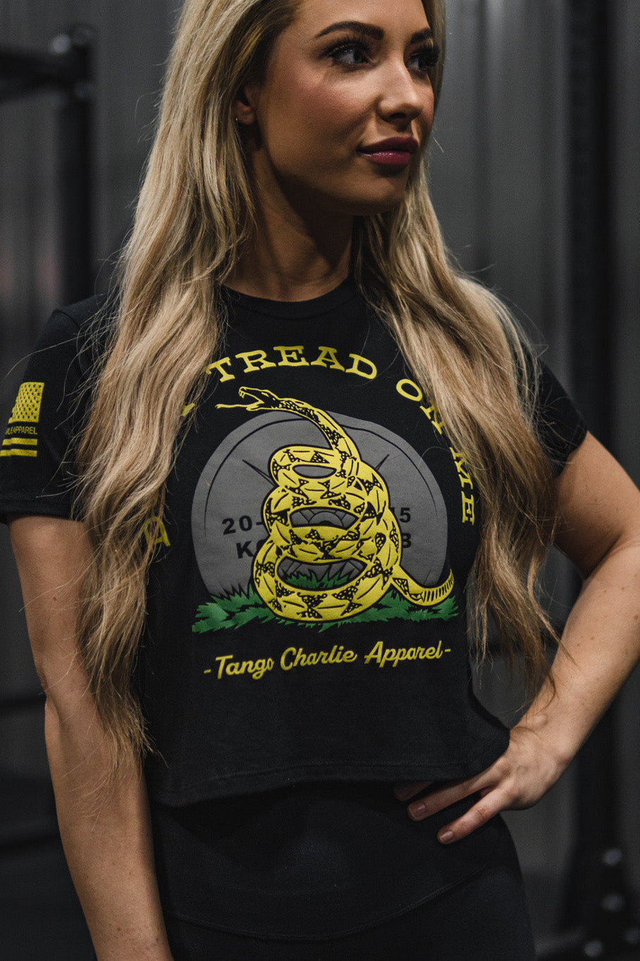 A woman stands indoors wearing the Don’t Tread On Me Crop Tee by Tango Charlie Apparel—a black crop top featuring a bold yellow snake graphic and slogan, crafted from soft polyester-cotton blend by Next Level for superior comfort and style.
