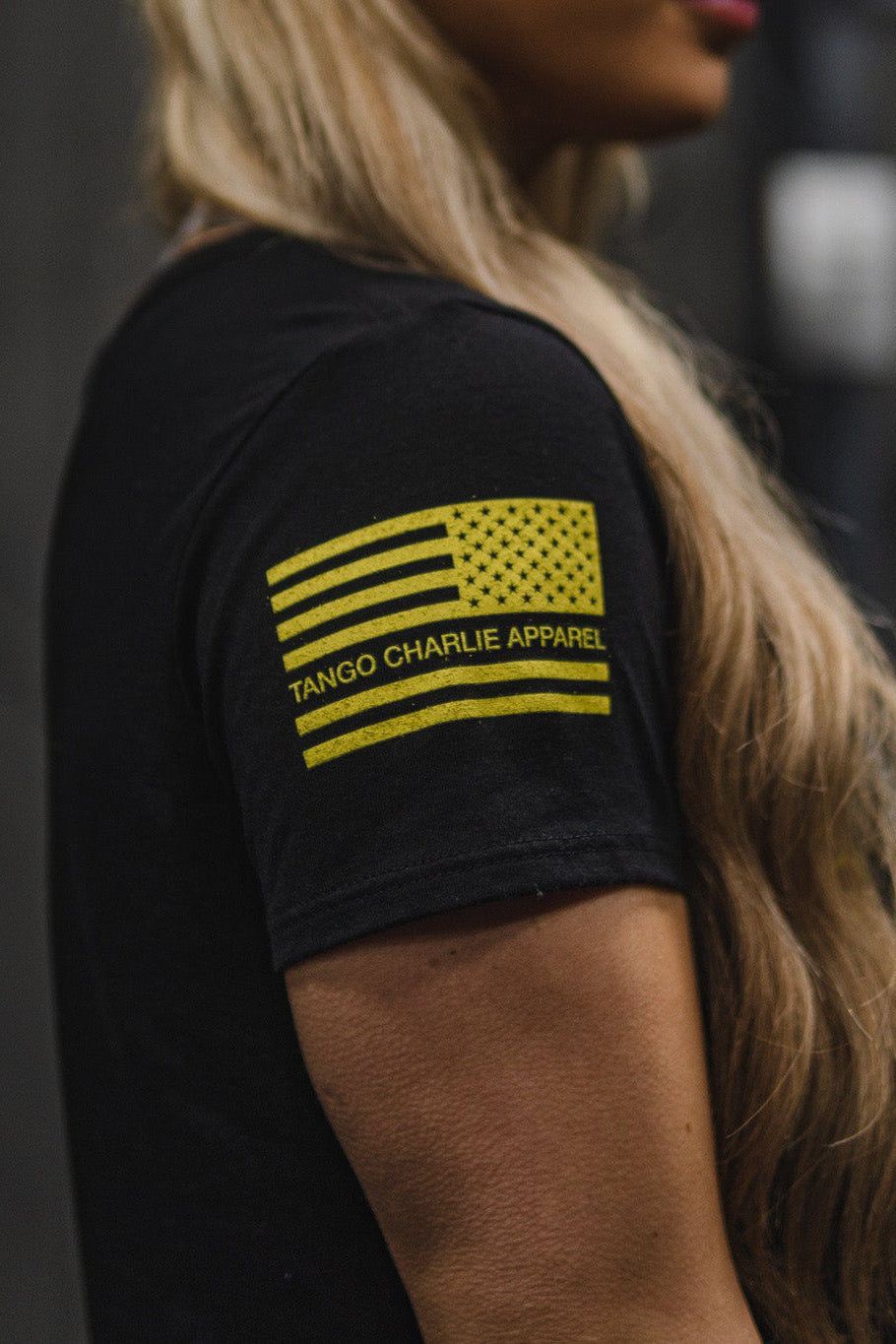 A person is shown wearing the Tango Charlie Apparel Don’t Tread On Me Crop Tee, a black polyester-cotton T-shirt featuring a yellow American flag graphic. Only their arm, shoulder, and part of their face are visible, subtly referencing iconic “Don’t Tread On Me” symbolism and highlighting next-level brand quality.