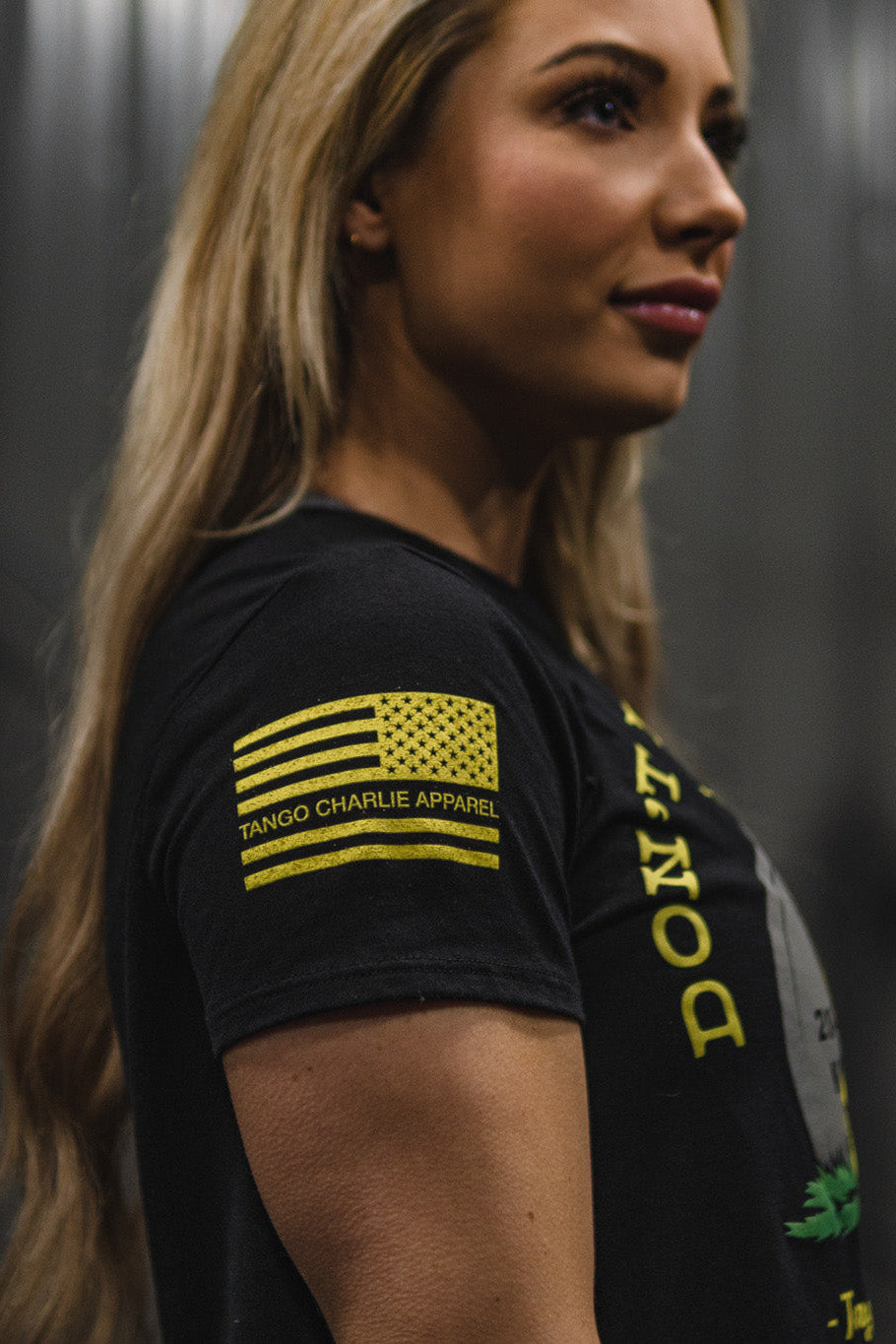 A woman with long blonde hair wears the “Don’t Tread On Me Crop Tee” from Tango Charlie Apparel, featuring a yellow American flag and Tango Charlie Apparel text on the sleeve. The black crop tee is crafted from a soft polyester-cotton blend for all-day comfort.
