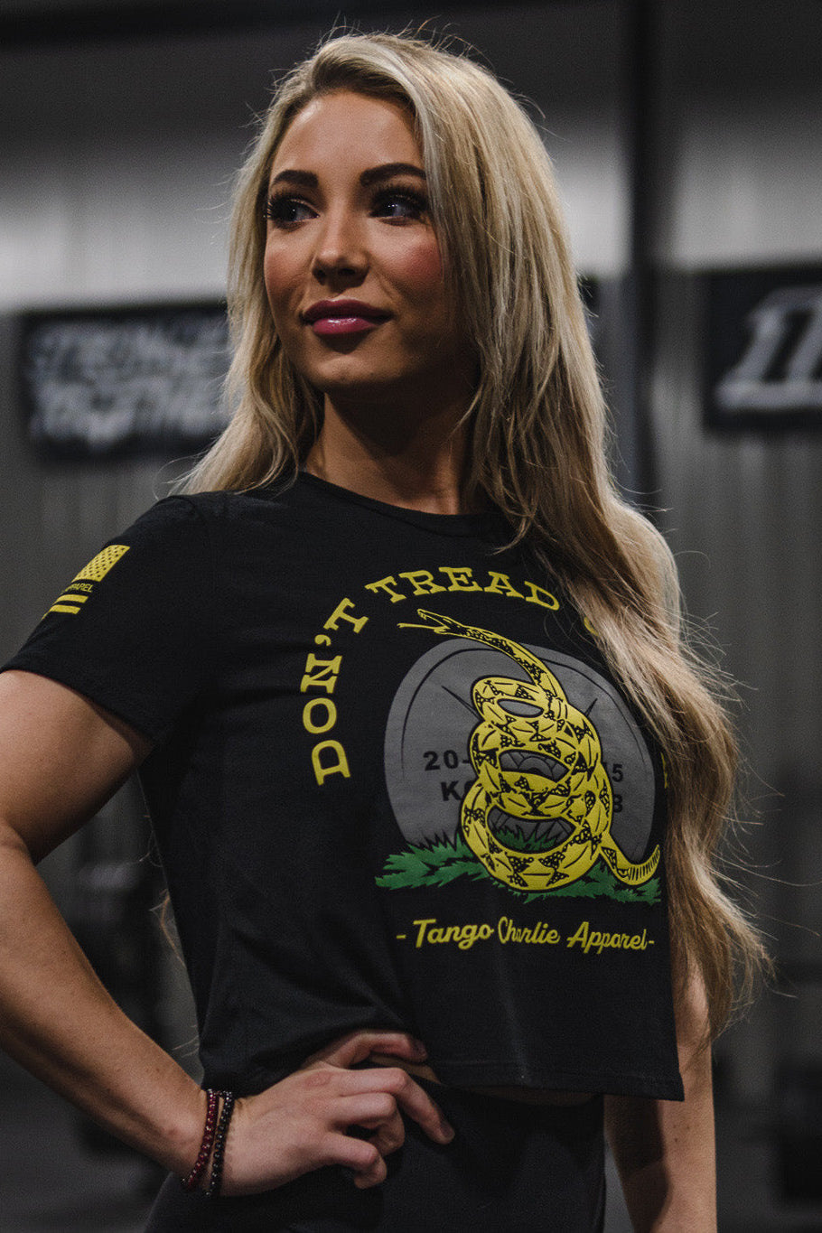 A woman with long blonde hair, wearing a black Don’t Tread On Me Crop Tee by Tango Charlie Apparel, stands indoors looking to the side with a neutral expression. The tee, made from a soft polyester cotton blend, offers both comfort and style as gym equipment appears blurred in the background.