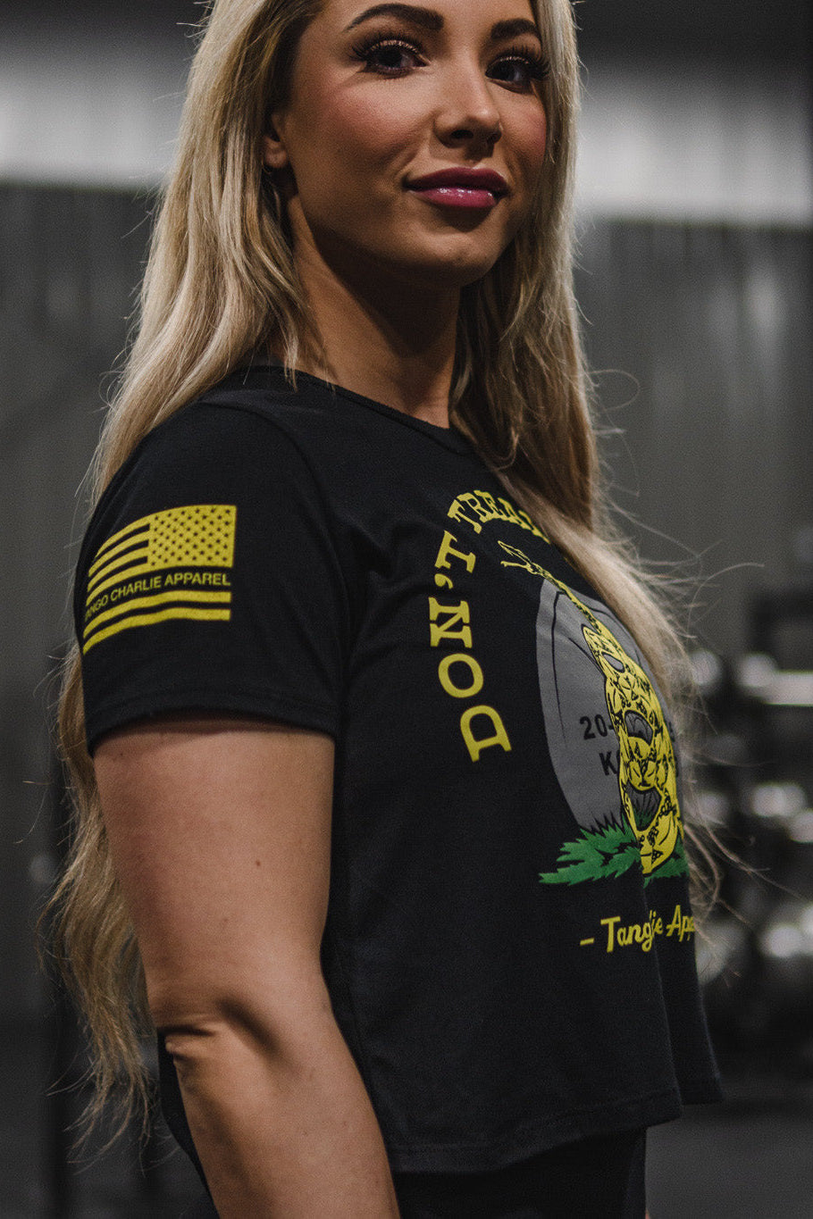 A woman with long blonde hair is wearing a black Don’t Tread On Me Crop Tee by Tango Charlie Apparel, made from soft polyester cotton, standing indoors in a gym setting with blurred exercise equipment in the background.