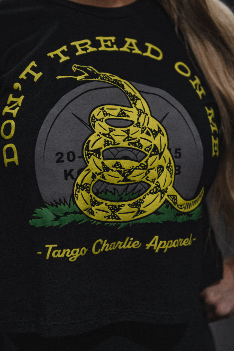 A person wearing the Don’t Tread On Me Crop Tee by Tango Charlie Apparel—a black, soft polyester-cotton shirt featuring a yellow coiled snake graphic and the text “DONT TREAD ON ME.”.