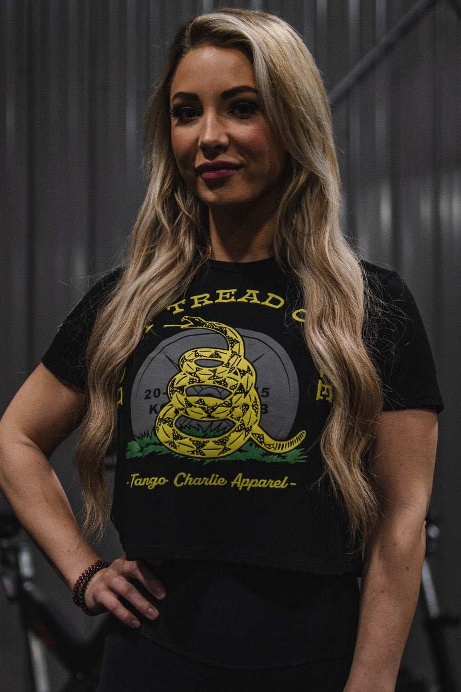 A woman with long blonde hair stands indoors wearing the Tango Charlie Apparel Don’t Tread On Me Crop Tee, a black top crafted from a soft polyester cotton blend and featuring a yellow coiled snake graphic with “Don’t Tread On Me” text.