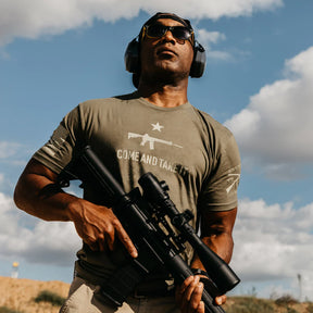 A man wearing military green earmuffs, sunglasses, and a Come And Take It t-shirt from Grunt Style stands with a rifle. The shirt features an image of a rifle, embodying the 2A Edition spirit against a backdrop of partly cloudy skies, expressing the right to bear arms.