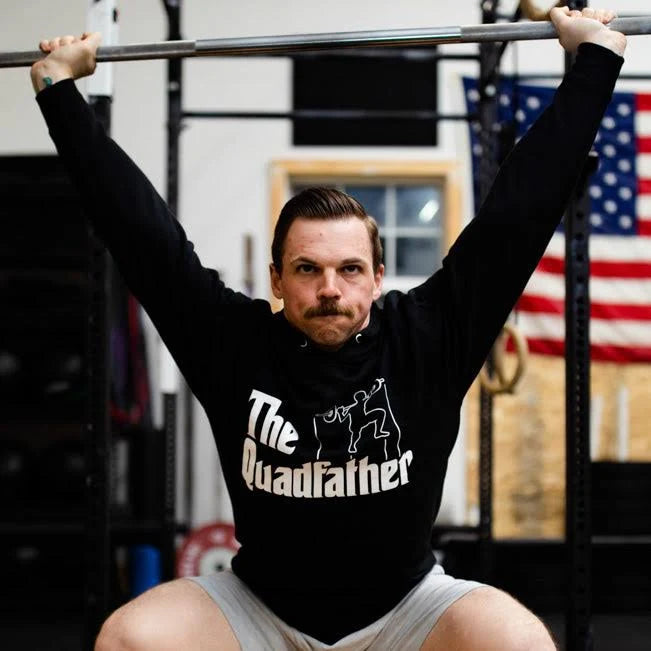 A man in a gym hoists a barbell overhead, wearing Tango Charlie Apparels The Quadfather Hoodie. An American flag in the background adds to the scenes intensity and pride.